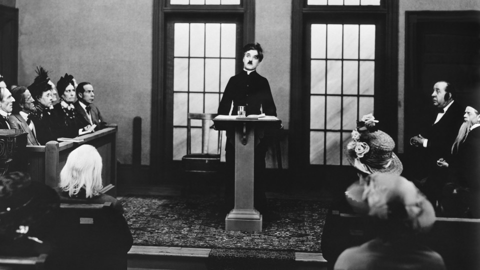 A black-and-white photo of Charlie Chaplin at a podium, surrounded by onlookers in pews