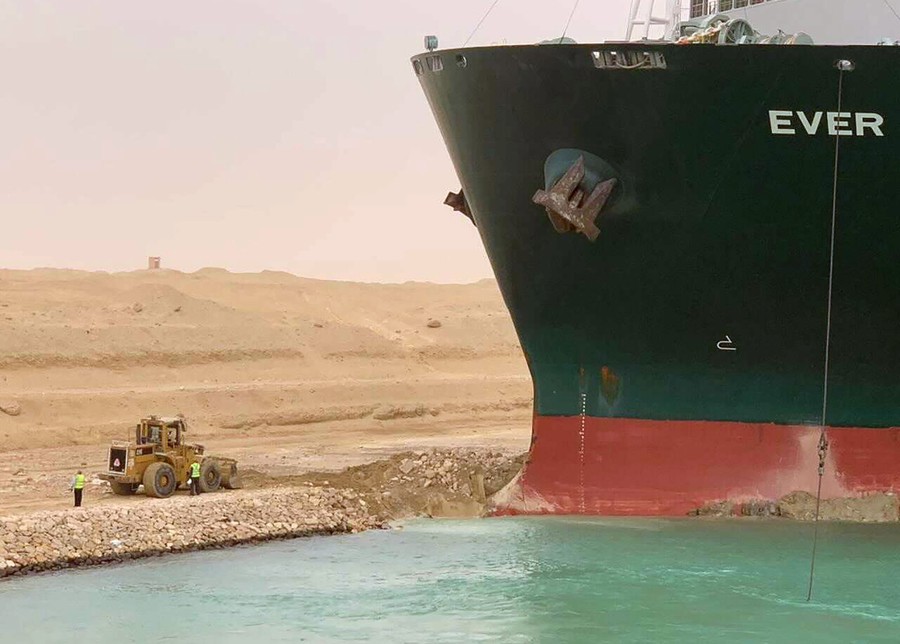 A large ship is seen with its front end jammed up against the sandy banks of a canal, with a relatively tiny front loader on the shore nearby.