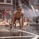 Children play in a city in front of rowhouses, in their swimwear, as a fire hydrant sprays water. One boy, front and center, seems excited and grins directly at the camera.