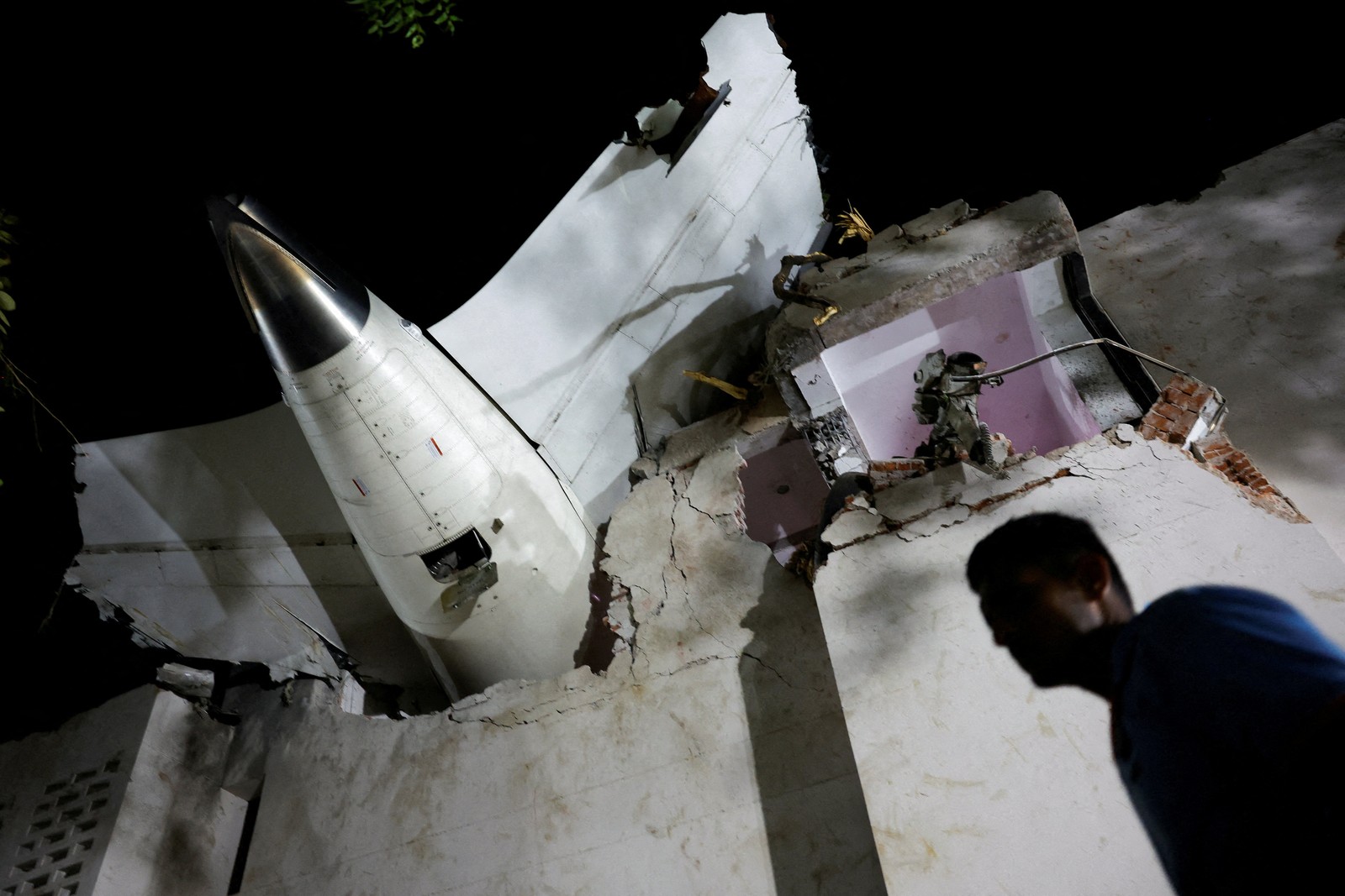 A person walks past debris at a crash site, beneath the tail section of an aircraft resting on top of a damaged building.