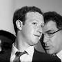 A black and white photograph of a bespectacled man whispering into Mark Zuckerberg's ear