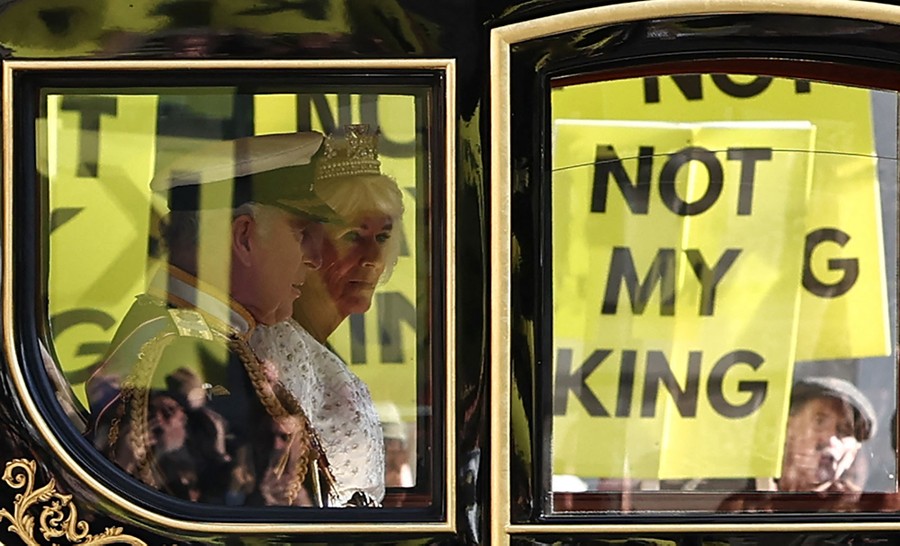 Protestors holding signs that read "Not My King" are captured through the windows of a carriage ridden by King Charles and Queen Camilla.