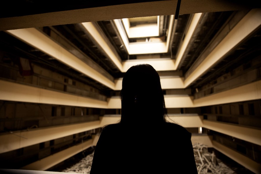 A silhouette of a woman is seen inside a dark hotel interior, a large and damaged space lit by a skylight above.