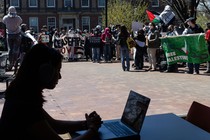 A college student works on her computer with pro-Palestinian protestors in the background.