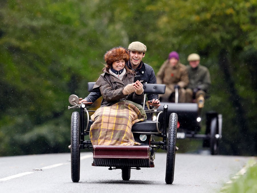 People laugh while driving down a road in a vintage three-wheeled vehicle.