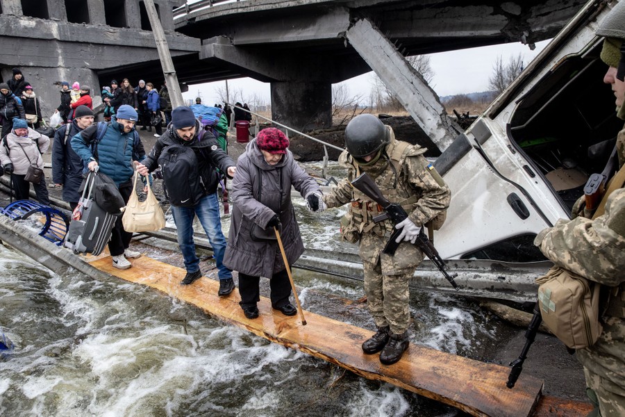 Soldiers help civilians walking on a broad plank across swiftly flowing water beneath a destroyed bridge.