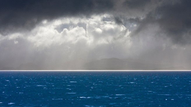An image of storm clouds with sun shining through onto a hyper-blue ocean