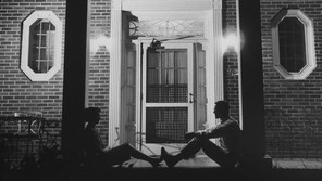 Black-and-white photo of a man and a woman sitting in front of a building at night. Each is leaning against opposite pillars, facing each other, with their feet touching.