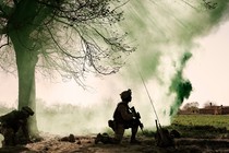 Picture of a Marine under a tree kneeling behind a smoke screen, holding a gun pointed toward the ground. Another Marine kneels behind the tree.