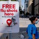 Photo of a woman on a city sidewalk wearing medical scrubs and a mask, next to a sign promoting free flu shots