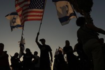 Israeli protesters are silhouetted against a darkening sky waving several Israeli flags and an American flag.