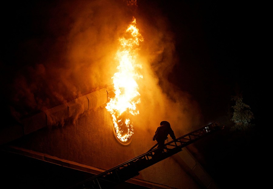 A firefighter climbs a ladder beside a burning building at night.
