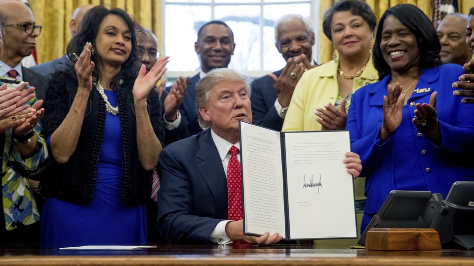 Donald Trump sits at his desk in the Oval Office holding a signed executive order. People smile and clap behind him.