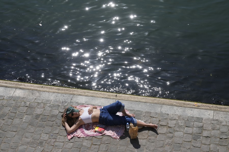 A woman lies on a towel on pavement beside a river, enjoying the sunshine.