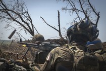 Several soldiers stand in a trench, as one fires a machine gun.