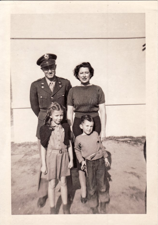 sepia-toned family photo of man in military uniform and woman with curled hair standing behind a young girl in dress and young boy in cuffed jeans, all squinting in bright sunlight