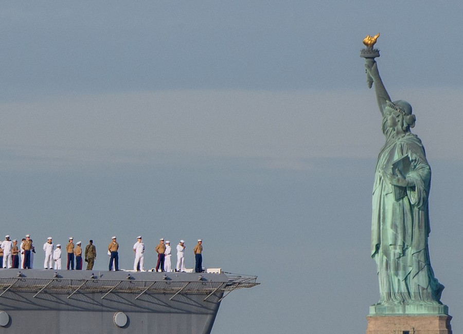 Sailors on a naval ship are seen passing by the Statue of Liberty.