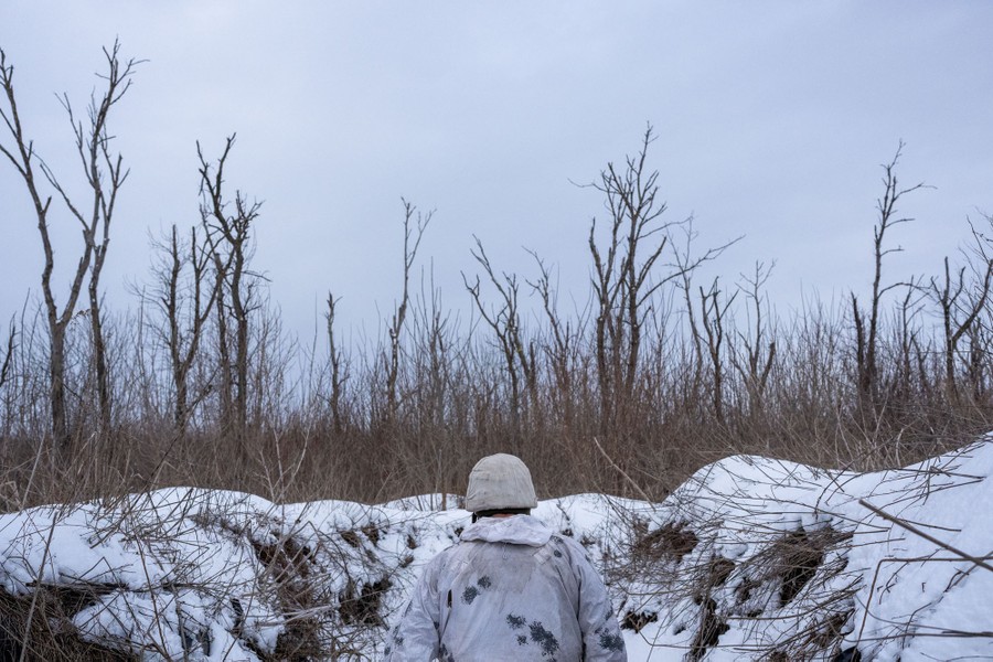 A soldier wearing white camouflage gear walks in a trench in a snow-covered field among leafless trees.