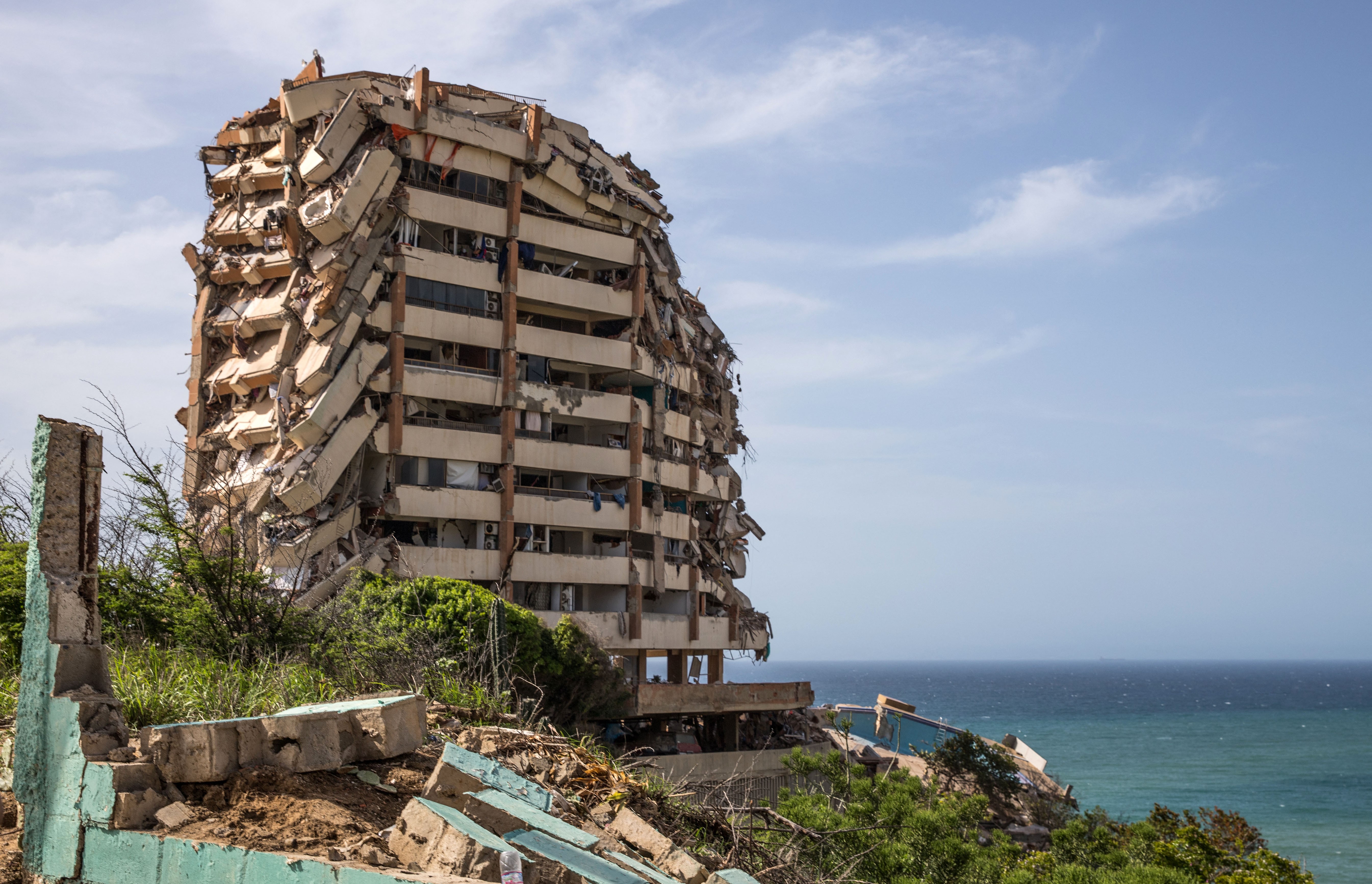 A heavily-damaged multi-story building, partially collapsed, seen along a shoreline.