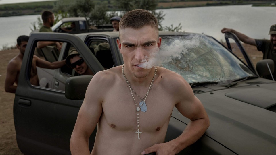 Ukrainian soldiers rest by a car; a shirtless man wearing a rosary stares at the camera while exhaling smoke.