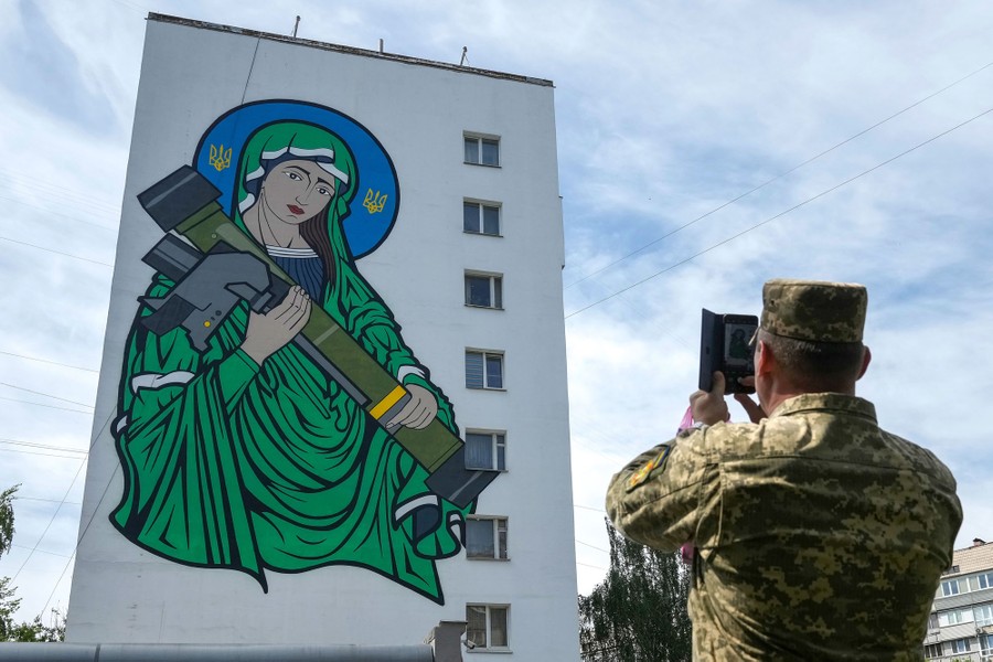 A soldier takes a photo of a large mural on the side of a building depicting a woman cradling a small missile launcher.