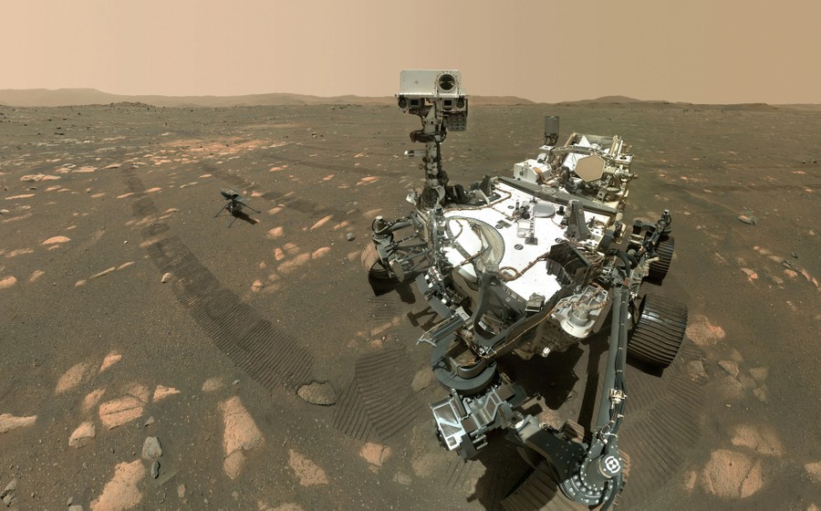 A large robotic rover is seen on Mars, with a tiny helicopter on the ground beside it.