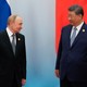 Xi Jinping holds his arm out while looking at Vladimir Putin, in front of flags of China and Russia.