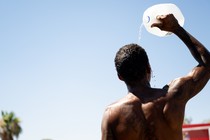 A shirtless man seen from the back standing in the sun, pouring a jug of water over his head