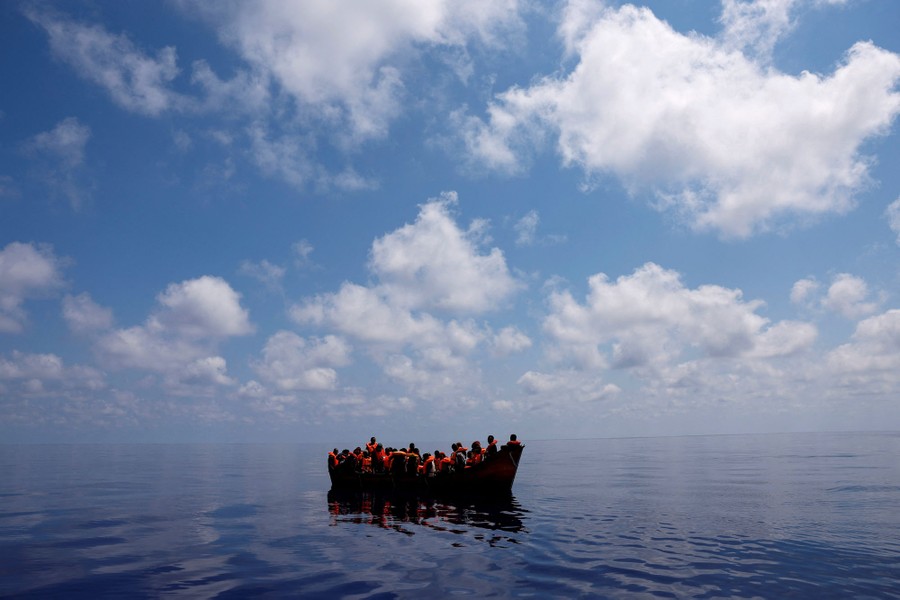 A couple dozen people wearing life vests ride in a small boat in calm water in a wide area of ocean.