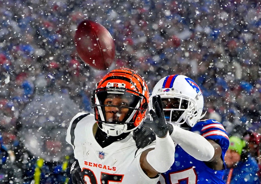 Two football players reach out for a football during a snowstorm.