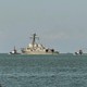 Two people in the foreground look out at a U.S. Navy warship in the water at a distance.