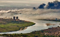 An elevated view of a river running through a city with black and white smoke rising from numerous fires.