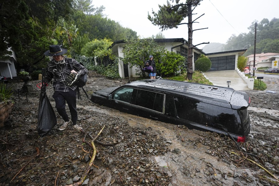 People carry items from a house, past a vehicle that sits half-buried in the debris of a mudslide.