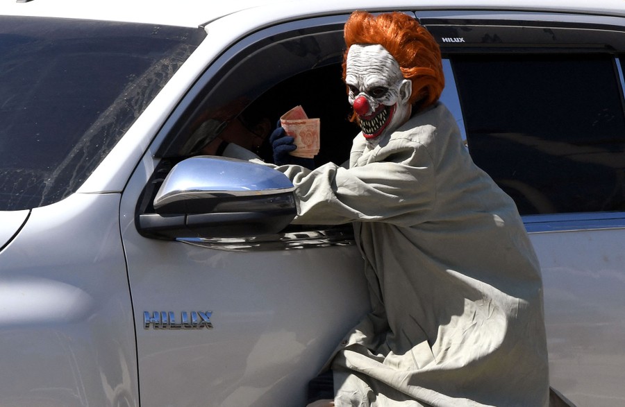 A person wears a frightening clown mask and long coat, reaching into the window of a passing vehicle, apparently asking for money.