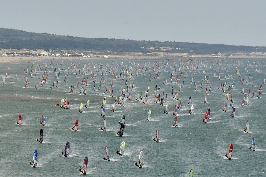 Hundreds of windsurfers hurtle across the water's surface near a beach.