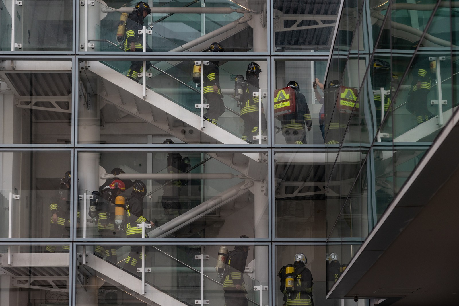 Firefighters in full gear climb the stairs of a tall building, seen from outside through a glass wall.