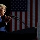 Donald Trump stands at a lectern in front of a flag. He is spotlighted; the rest of the scene is dark.