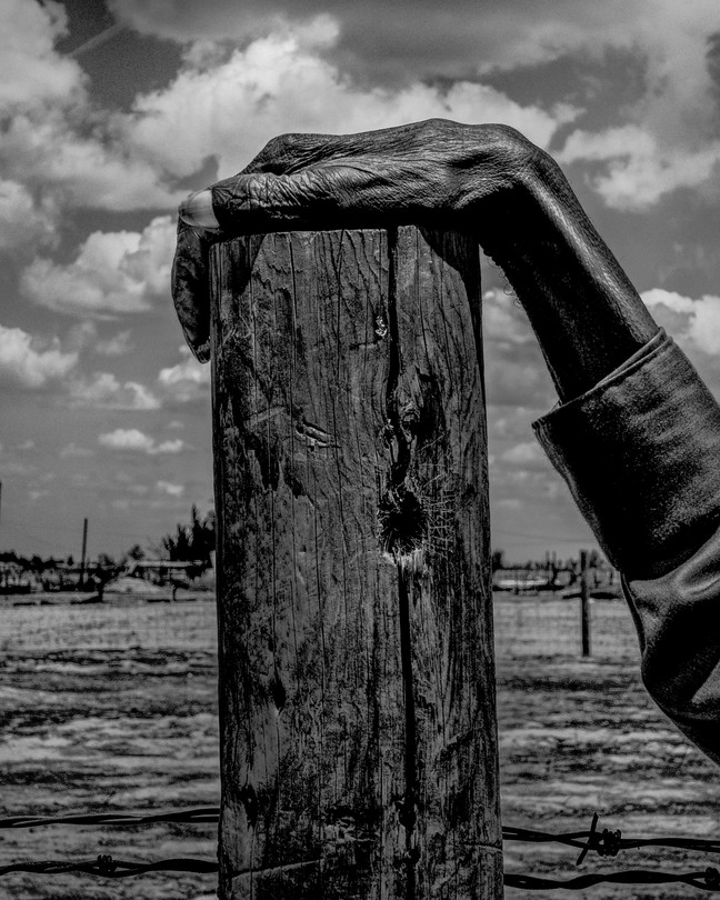 An old man's hand rests on a post in a drought landscape in central California