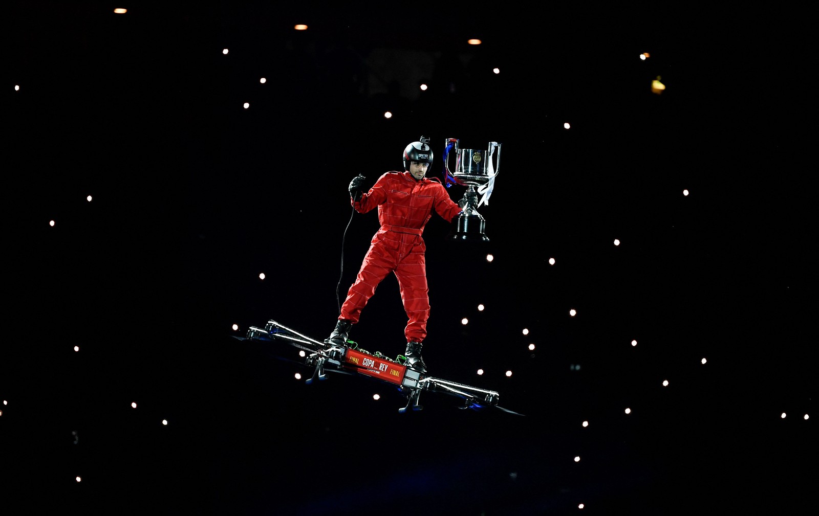 A person holds a trophy while standing atop a large rideable quadcopter, during a ceremony.