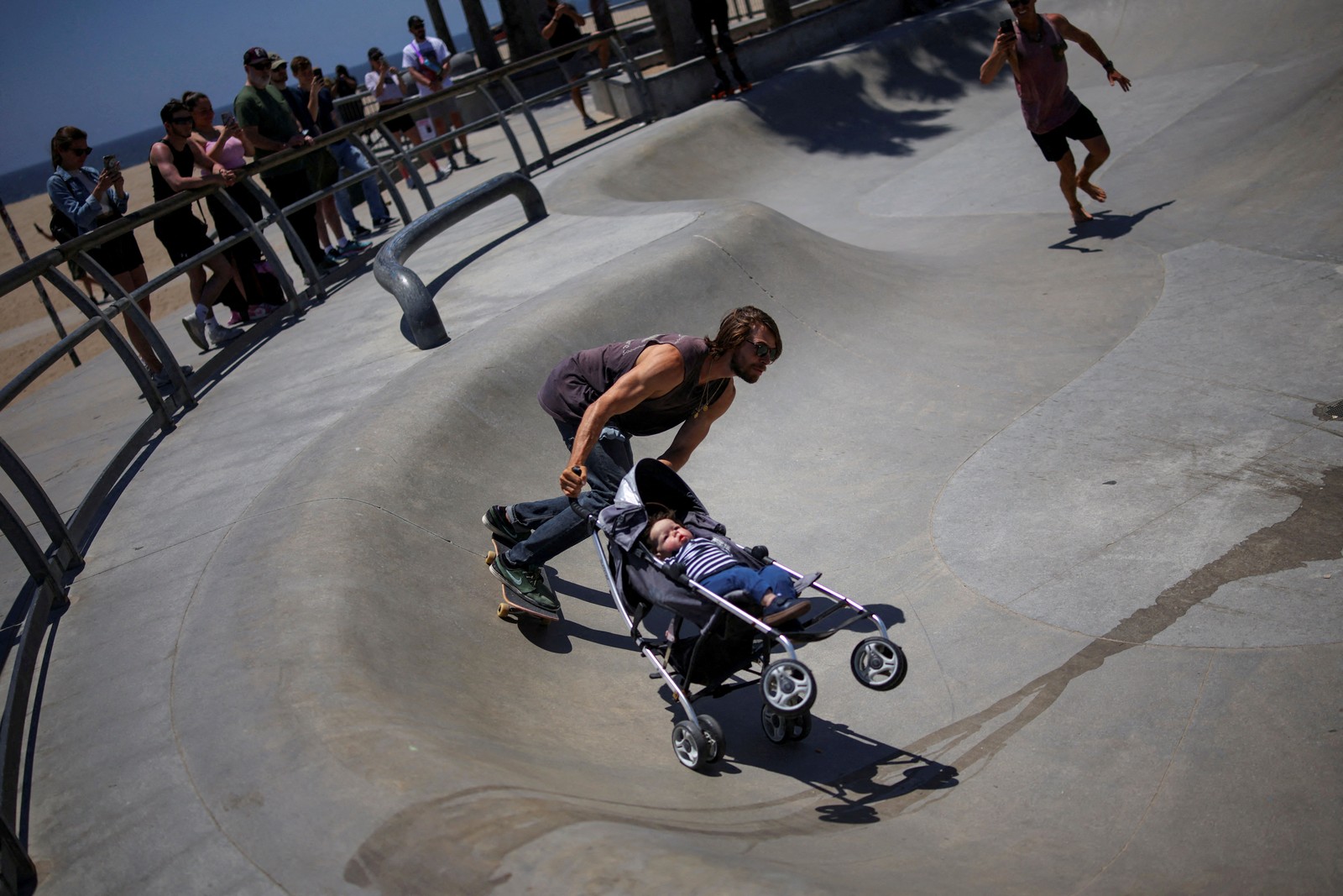 A skateboarder pushes a stroller with a doll as he rides through a skate park.