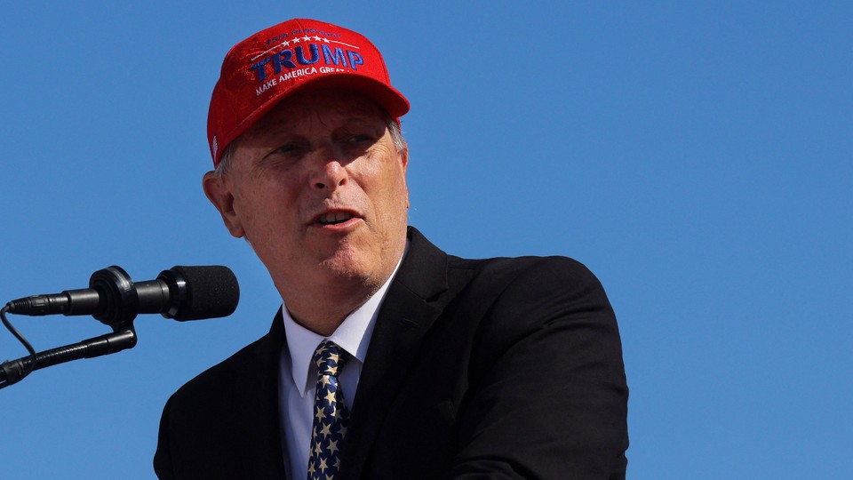 Andy Biggs stands before a microphone wearing a "Trump: Make America Great Again" hat.