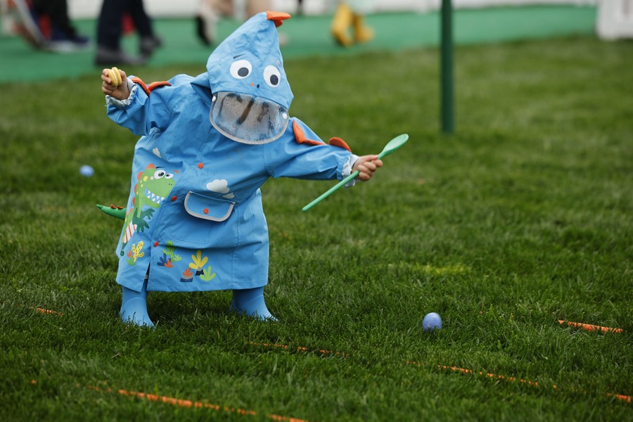 A child wearing a playful raincoat decorated with eyes on the hood leans over to nab an Easter egg on grass.