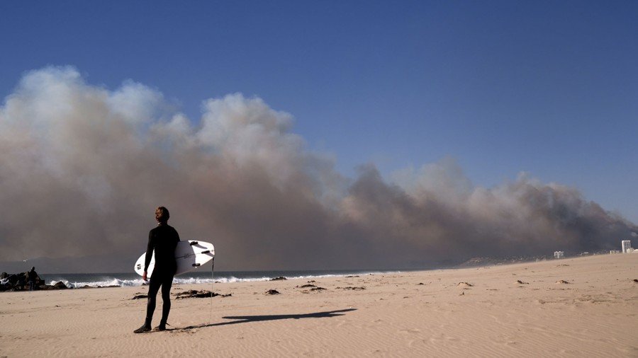 A surfer on a beach holds a surf board and looks at dark smoke along the horizon from a distant fire.