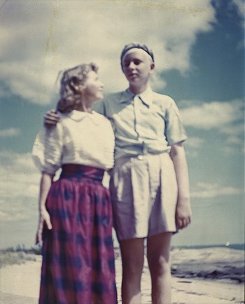 photo: a woman in white shirt and plaid skirt looks up at her taller son, wearing shorts, with blue sky and beach in background