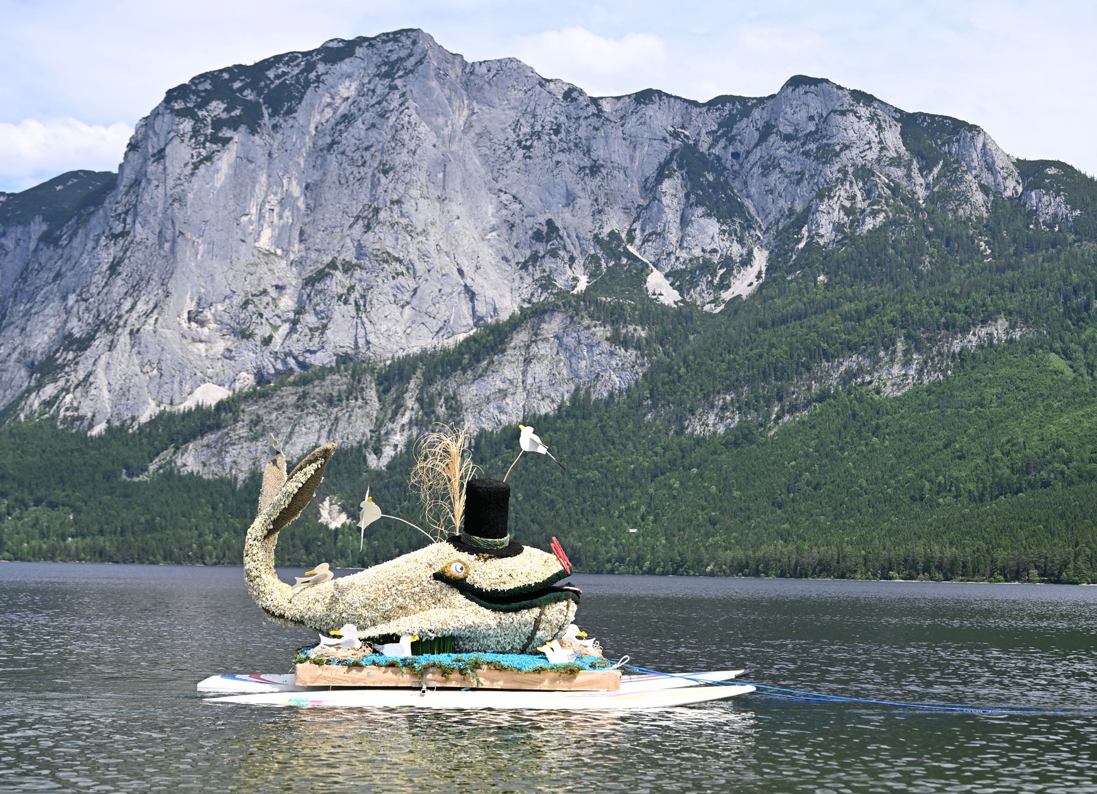 A large whale sculpture made of daffodils floats in a lake near tall mountains.