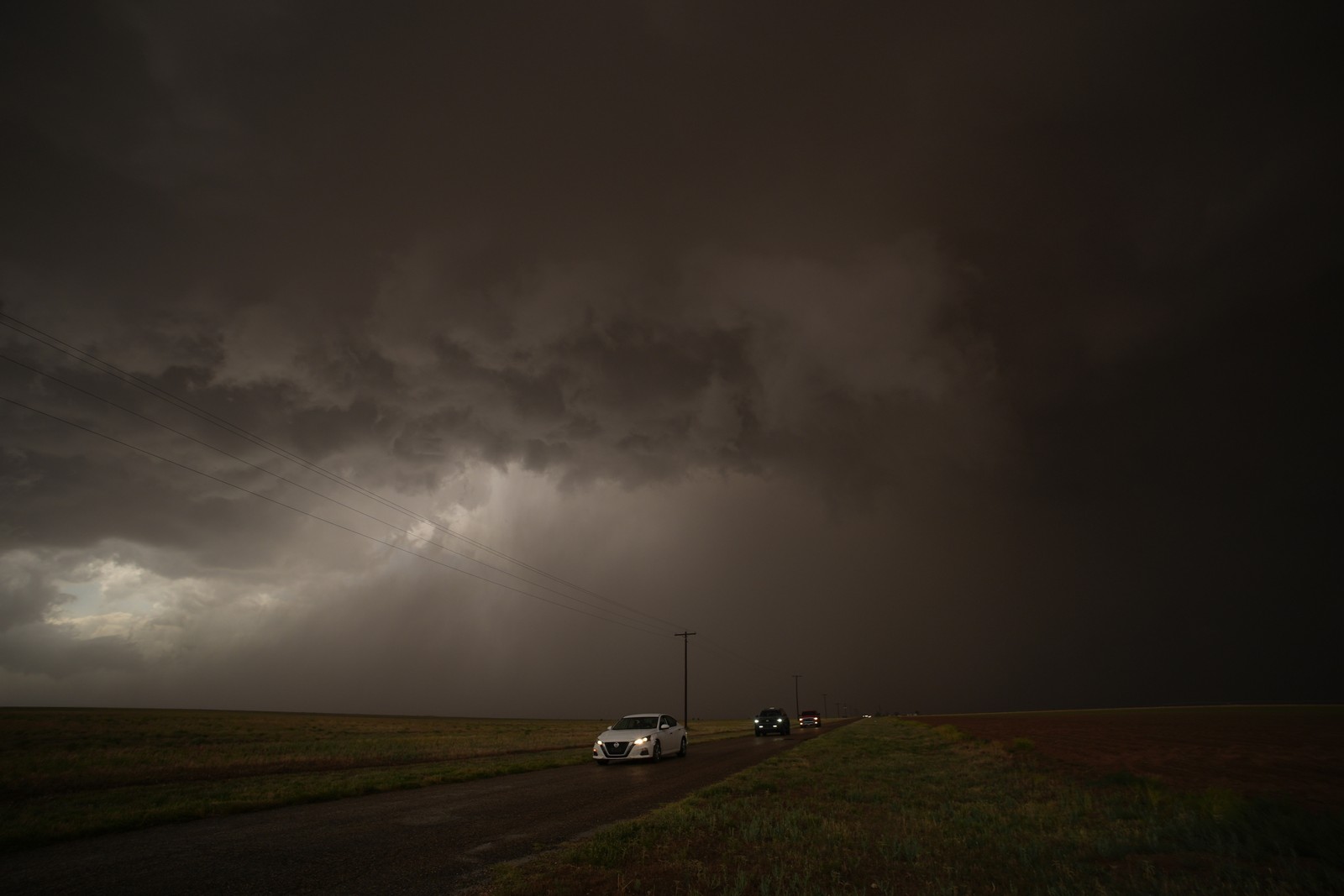 Several cars drive on a road through wide-open farm country, beneath very dark clouds.