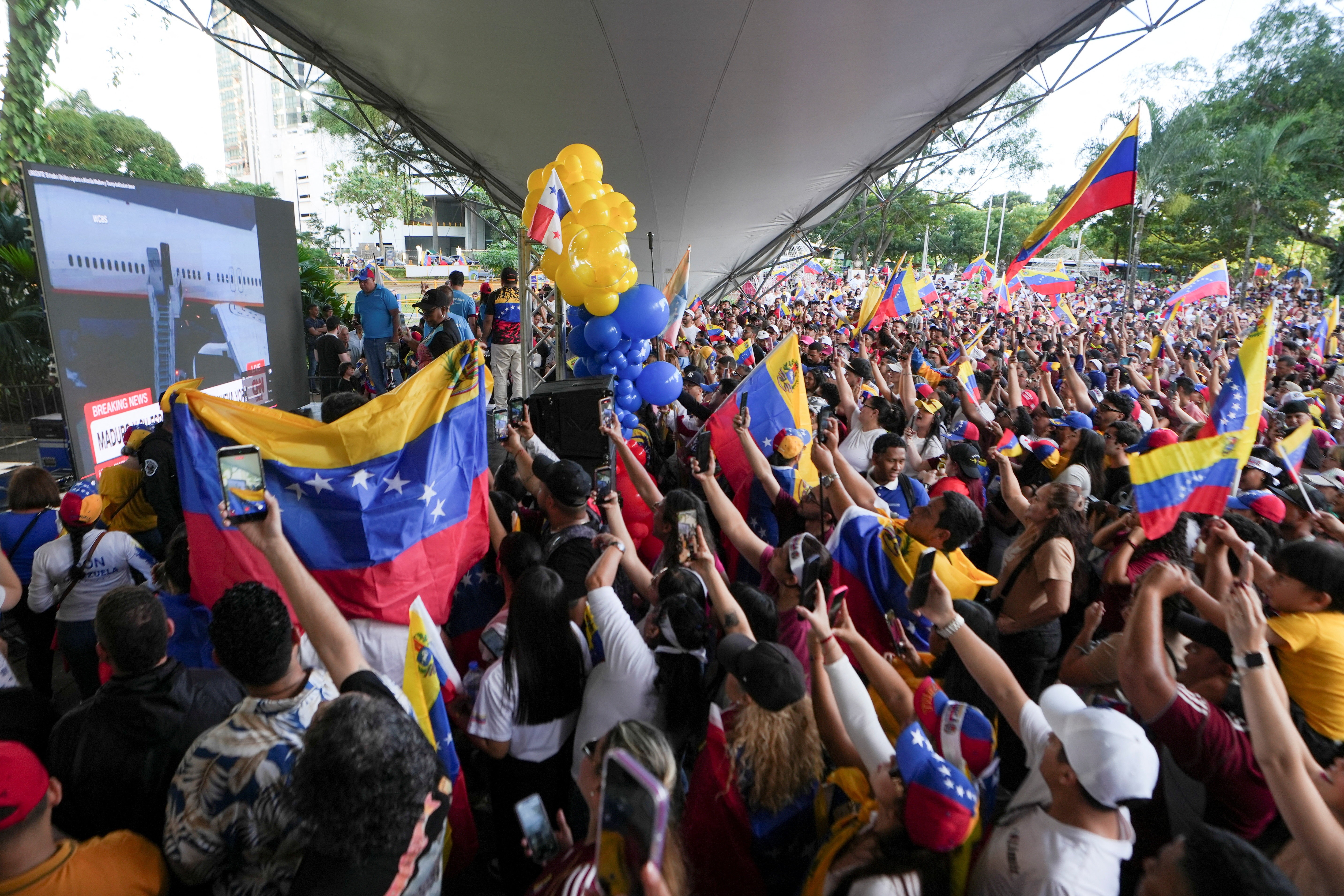 A crowd of people holding Venezuelan flags cheer while watching a CNN news broadcast.