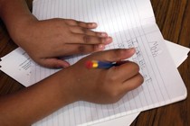 A child holds a pencil and writes on a sheet of notebook paper. 