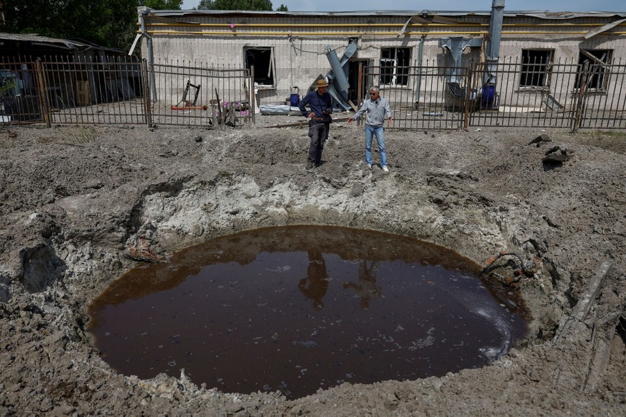 Two people stand beside a water-filled bomb crater, next to damaged buildings.