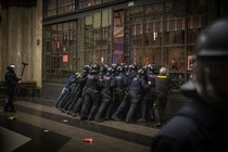 A group of riot police pushes together against a large door with protesters on the other side.